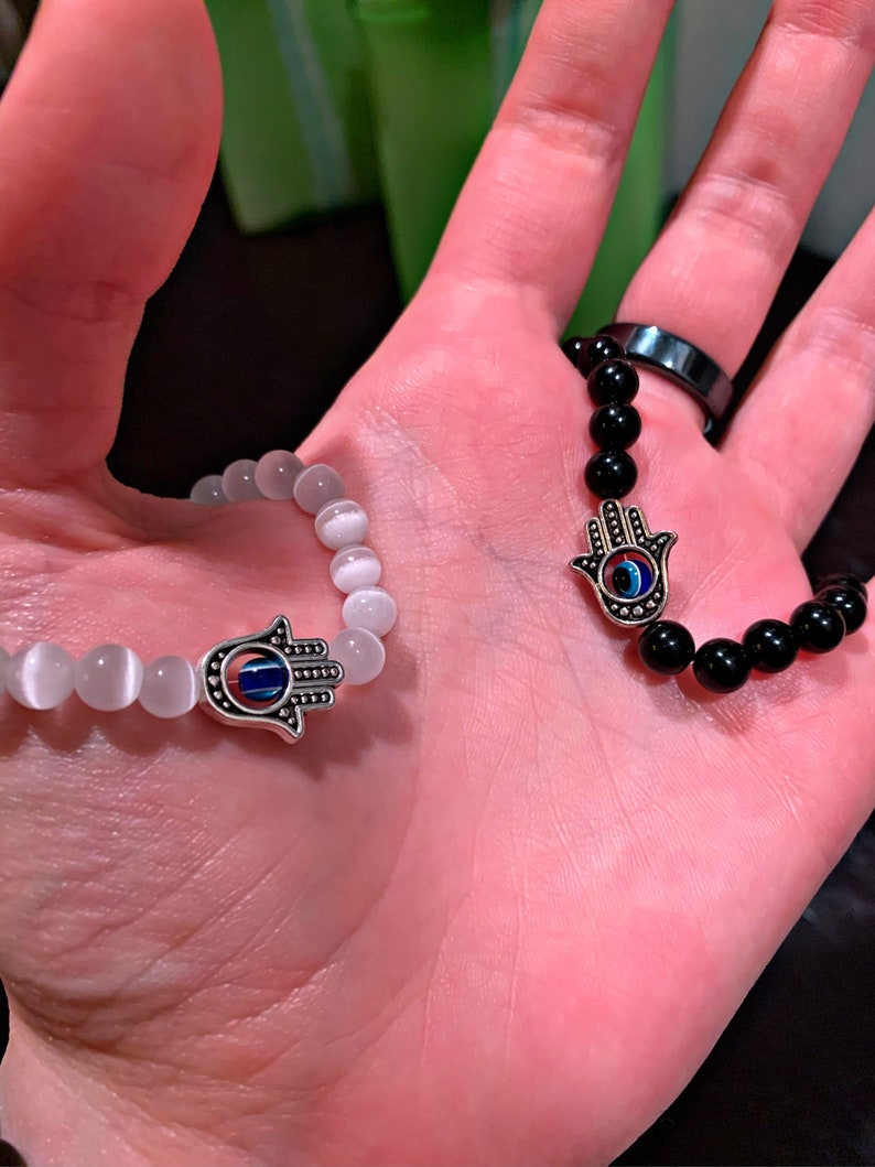 Puede incluir: Dos pulseras con dijes de mano de hamsa. Una pulsera tiene cuentas blancas y la otra tiene cuentas negras. Ambos dijes tienen un ojo turco azul en el centro.