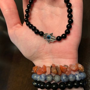 Puede incluir: Un brazalete de cuentas negras con un dije de mano de hamsa plateado con un ojo turco azul. El brazalete se lleva en la mu&ntilde;eca con otros brazaletes.