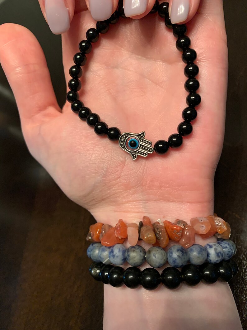 Puede incluir: Un brazalete de cuentas negras con un dije de mano de hamsa plateado con un ojo turco azul. El brazalete se lleva en la mu&ntilde;eca con otros brazaletes.