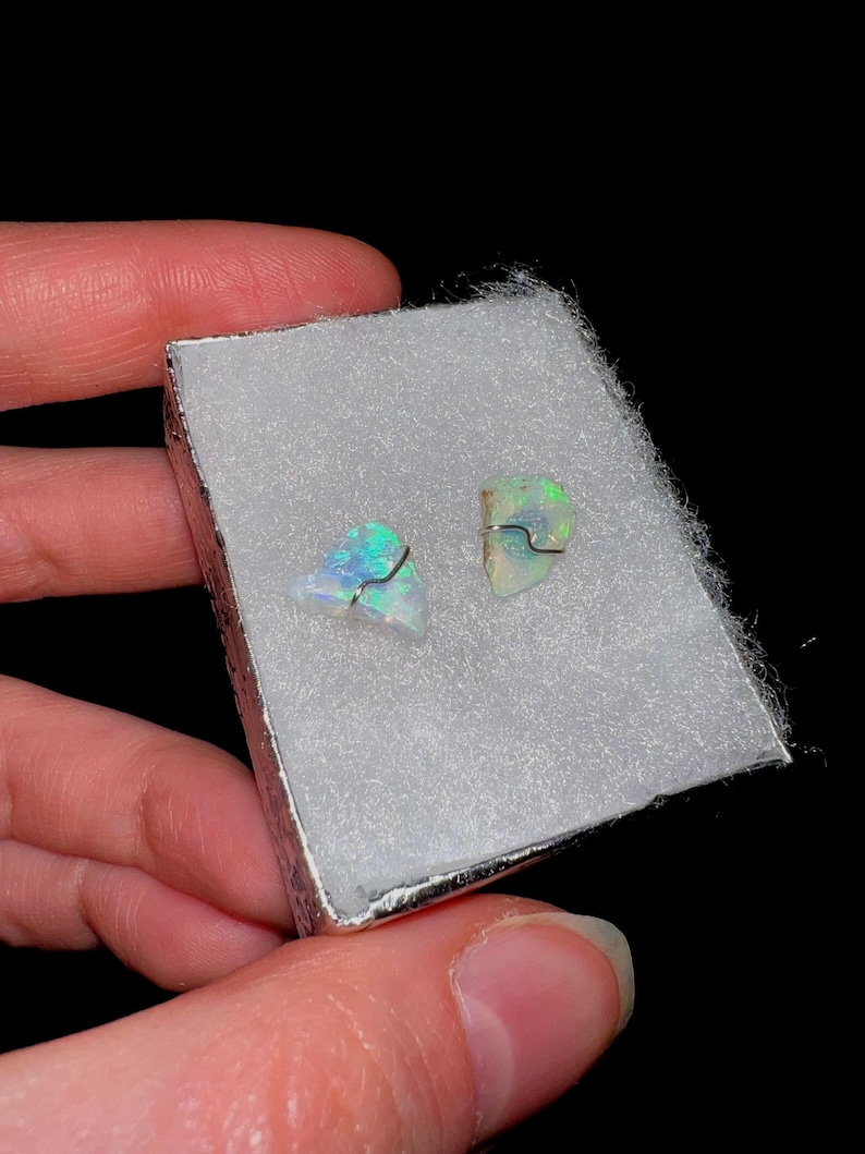 May include: Two small, iridescent opal gemstones set in silver wire, displayed in a white box with a white velvet lining.