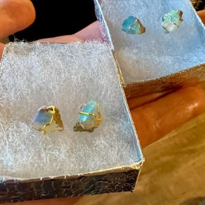 May include: Two pairs of earrings with blue and green iridescent stones wrapped in gold wire. The earrings are displayed in two small white boxes with a textured silver exterior.