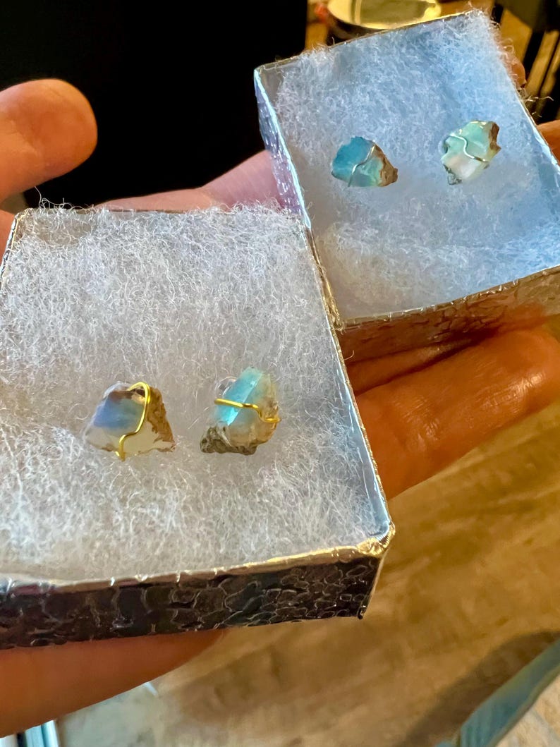 May include: Two pairs of earrings with blue and green iridescent stones wrapped in gold wire. The earrings are displayed in two small white boxes with a textured silver exterior.