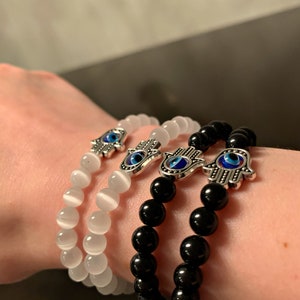 Puede incluir: Tres pulseras con dijes de mano de hamsa plateados con detalles de ojo turco azul. Una pulsera est&aacute; hecha con cuentas blancas, una con cuentas negras y una con cuentas blancas.