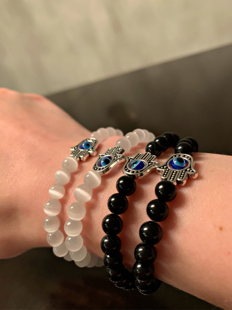 Puede incluir: Tres pulseras con dijes de mano de hamsa plateados con detalles de ojo turco azul. Una pulsera est&aacute; hecha con cuentas blancas, una con cuentas negras y una con cuentas blancas.
