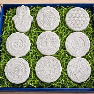 Könnte beinhalten: Ein Set aus neun weißen, runden und hamsa-förmigen Plaketten mit detaillierten geprägten geometrischen und spirituellen Symbolen, darunter der Lebensbaum, Om und die Blume des Lebens, in einer blauen Schachtel mit grünem Papierfüllmaterial.