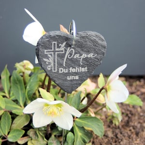 May include: A heart-shaped slate stone with the words "Papa Du fehlst uns" engraved on it. The stone is attached to a metal stake and is surrounded by white flowers.