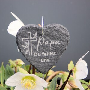 May include: A gray heart-shaped slate garden stake with a white cross and the words "Papa Du fehlst uns" engraved on it. The stake is placed in a pot of white flowers.