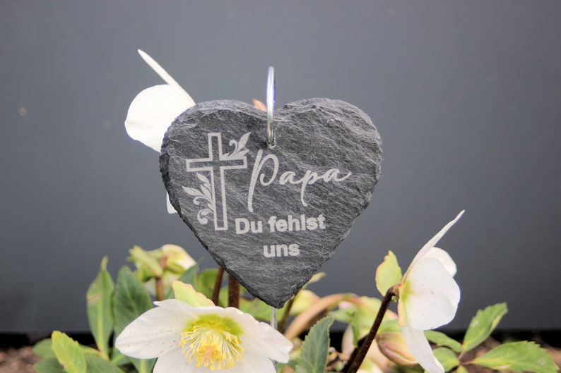 May include: A gray heart-shaped slate garden stake with a white cross and the words "Papa Du fehlst uns" engraved on it. The stake is placed in a pot of white flowers.