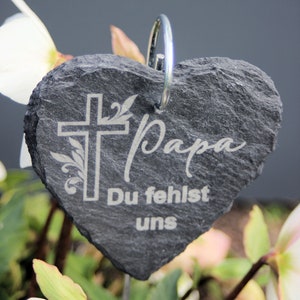 May include: A gray heart-shaped slate stone with a silver ring at the top. The stone has a cross with a floral design and the words "Papa Du fehlst uns" engraved on it.