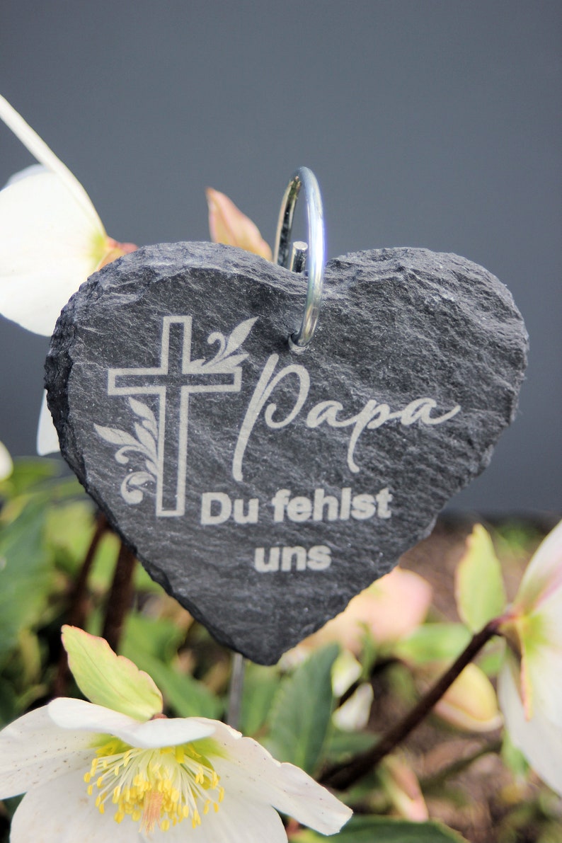 May include: A gray heart-shaped slate stone with a silver ring at the top. The stone has a cross with a floral design and the words "Papa Du fehlst uns" engraved on it.