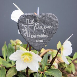 May include: A black heart-shaped slate plaque with the words "Papa Du fehlst uns" and a cross engraved on it. The plaque is attached to a metal stake and placed in a bed of white flowers.