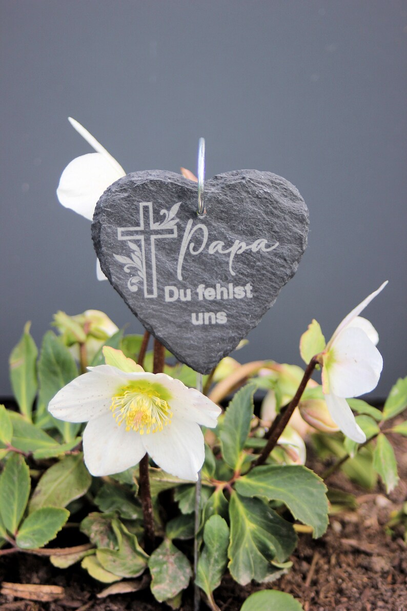 May include: A black heart-shaped slate plaque with the words "Papa Du fehlst uns" and a cross engraved on it. The plaque is attached to a metal stake and placed in a bed of white flowers.