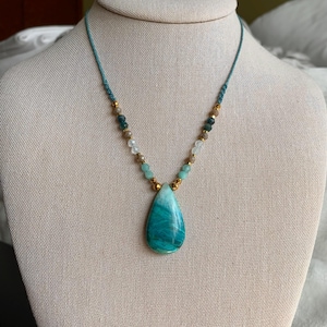 May include: A teal and gold beaded necklace with a large, teardrop-shaped pendant. The pendant is a marbled teal color. The necklace is displayed on a mannequin. The necklace is a piece of jewelry.