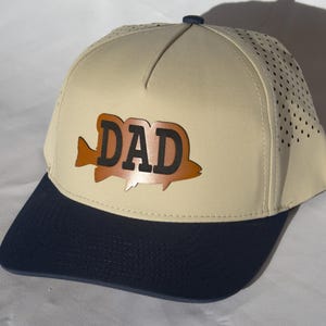 May include: A beige baseball cap with a dark blue brim and a dark blue button on top. The front panel features a brown fish-shaped patch with the word DAD printed in bold black letters. The side panels of the cap have a perforated design.
