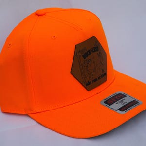 May include: A bright neon orange baseball cap featuring a brown hexagonal leather patch on the front with a black graphic illustration and the text "THE DUCK-LESS BUT FUN AT CAMP". A rectangular sticker is attached to the flat brim of the cap.