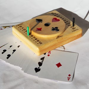 May include: A wooden card game scorekeeper with a circular rotating dial featuring four suit symbols. The board includes two rows of numbers from 0 to 10, labeled Team 1 and Team 2, with a green peg and a black peg inserted. A deck of playing cards is fanned out underneath.