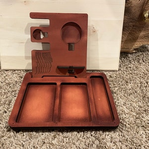 Op de afbeelding: Een houten valetstandaard met een dienblad en een standaard om spullen op te bergen. Het dienblad heeft drie vakken en de standaard heeft een klein vak voor een horloge, een groter vak voor een telefoon en een klein vak voor een ring. De standaard is versierd met een Amerikaanse vlag.