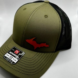 May include: A green and black trucker hat with a brown leather patch shaped like the Upper Peninsula of Michigan. The patch is orange in color. The hat has an adjustable strap and a label that reads "Est. Richardson 112 Original Trucker Adjustable".