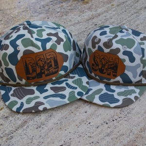 May include: Two baseball caps featuring a camouflage pattern in shades of green, brown, and white. Each cap has a brown leather patch on the front. The larger cap displays the word DAD, and the smaller cap displays the word SON, both with mountain and deer silhouettes.