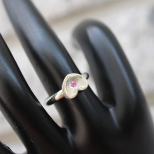 May include: A silver ring with a heart-shaped design. The heart features a small, round, pink gemstone at its center. The ring is displayed on a black surface, with a white background visible in the distance. Jewelry.