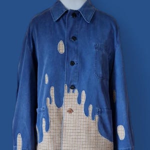 Könnte beinhalten: Eine blaue Jeansjacke mit einer beigen und braunen Hahnentritt-Applikation am unteren Saum sowie verstreuten ovalen Flicken an den Ärmeln und auf der Brust. Die Jacke verfügt über einen Kragen, eine Knopfleiste und zwei Seitentaschen.
