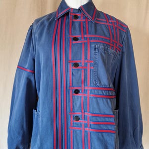 Könnte beinhalten: Blaue Jacke mit roten Bandakzenten. Die Jacke hat einen Kragen, schwarze Knöpfe und zwei Vordertaschen. Rote Banddetails sind in vertikalen und horizontalen Mustern auf der Vorderseite und den Ärmeln angeordnet.