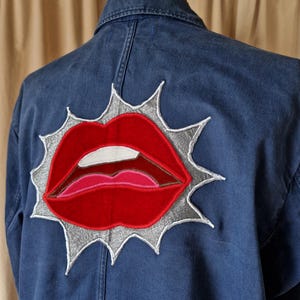 Könnte beinhalten: Dunkelblaue Jeansjacke mit einem großen, gestickten Design roter Lippen mit weißen und rosa Details, vor einem silbernen Burst. Die Jacke wird auf einer Schaufenquinpuppe präsentiert.