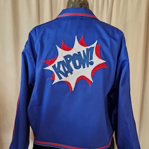 Könnte beinhalten: Blaue Jacke mit roten und weißen Akzenten. Auf der Rückseite befindet sich eine Comic-Grafik "KAPOW!" in Weiß mit roten und blauen Details. Die Jacke hat einen roten Streifen am Kragen und an den Manschetten.