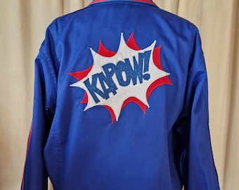 May include: A blue jacket with red and white accents. The back of the jacket features a comic book style "KAPOW!" graphic in white with red and blue details. The jacket has a red stripe along the collar and cuffs.