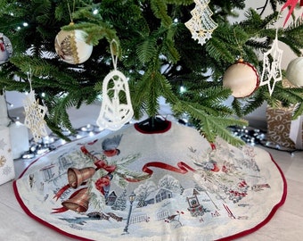 May include: A white and red Christmas tree skirt with a winter scene featuring a snowy village, a red ribbon, and two bells. The skirt has a red border.