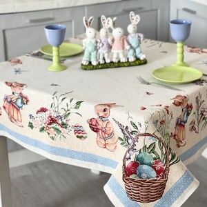Puede incluir: Un mantel blanco con un borde azul presenta un patr&oacute;n repetido de conejos, flores y huevos de Pascua. Una cesta llena de huevos de Pascua coloridos se encuentra en la esquina inferior derecha. Dos vasos azules y un plato verde est&aacute;n sobre la mesa.