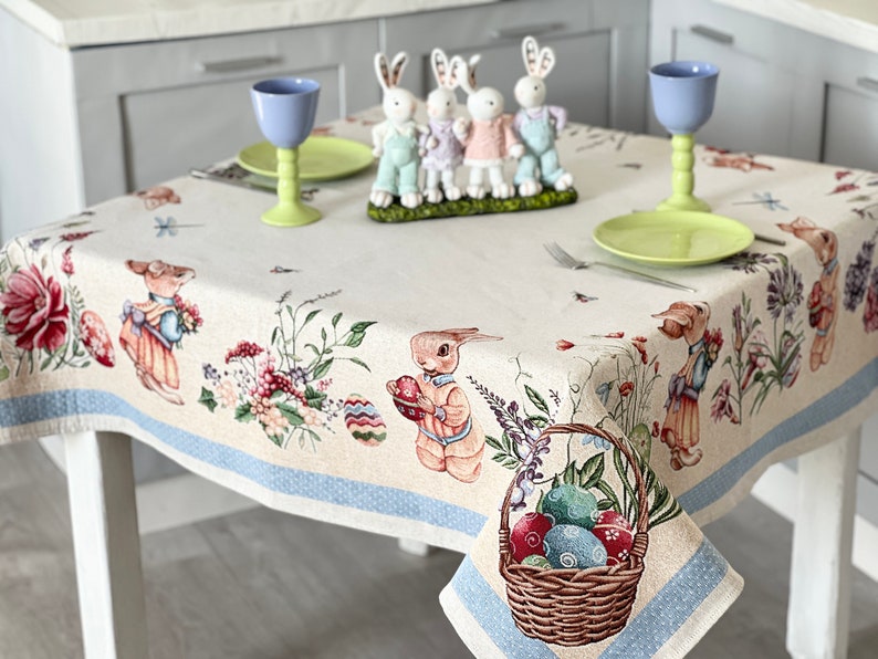 Puede incluir: Un mantel blanco con un borde azul presenta un patr&oacute;n repetido de conejos, flores y huevos de Pascua. Una cesta llena de huevos de Pascua coloridos se encuentra en la esquina inferior derecha. Dos vasos azules y un plato verde est&aacute;n sobre la mesa.
