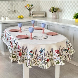 Könnte beinhalten: Ovale Tischdecke mit Blumen- und Vogelmuster. Die Tischdecke hat einen weißen Hintergrund mit einem Rand aus Blumen, darunter Gänseblümchen, Lavendel und rote Mohnblumen. Vier rosa Teller und vier blaue Gläser sind auf dem Tisch.