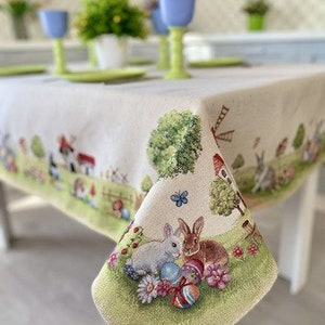 May include: A white tablecloth with a green, brown, and white Easter bunny and egg design. The tablecloth has a white background with a green border. The design features two bunnies holding Easter eggs, a windmill, and a small house.