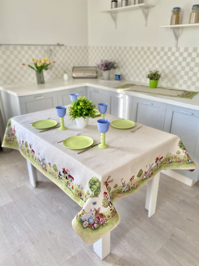May include: A white tablecloth with an Easter-themed design featuring bunnies, eggs, and a windmill. The tablecloth is set on a wooden table with four blue glasses and green plates.