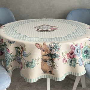 Puede incluir: Mantel redondo con un diseño caprichoso de conejito. La tela beige presenta una ilustración central de un conejito sosteniendo flores, rodeado de motivos florales y mariposas. El borde tiene un patrón de rayas azules y blancas, con un borde festoneado.