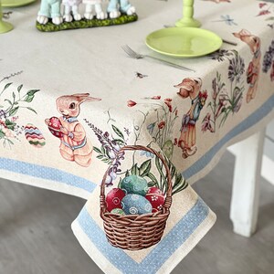 Puede incluir: Un mantel beige con un borde azul y un patr&oacute;n de lunares blancos. El mantel presenta una cesta de huevos de Pascua coloridos, conejos y dise&ntilde;os florales.