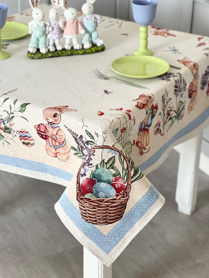 Puede incluir: Un mantel beige con un borde azul y un patr&oacute;n de lunares blancos. El mantel presenta una cesta de huevos de Pascua coloridos, conejos y dise&ntilde;os florales.