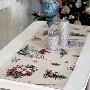 Puede incluir: Un camino de mesa blanco con una escena de invierno con esquiadores, árboles y ciervos. El camino de mesa tiene un fondo beige con acentos rojos, verdes y azules. Dos candelabros plateados están en el camino de mesa.