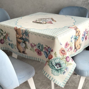 Può includere: Una tovaglia bianca con un bordo a quadri blu e bianchi presenta un motivo floreale con coniglietti che tengono dei fiori. La tovaglia è stesa su un tavolo con sedie blu.