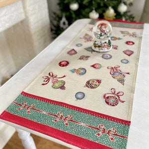 May include: A white table runner with a green and red border. The runner is decorated with a pattern of Christmas ornaments and bows. A small snow globe sits on the runner.