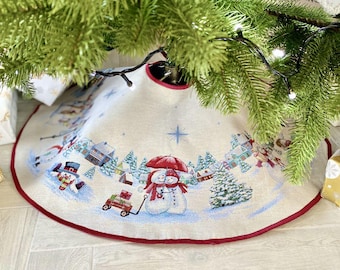 May include: A beige Christmas tree skirt with a red trim features a winter scene with two snowmen holding hands under a red umbrella. The scene also includes a red wagon with presents, houses, and trees.
