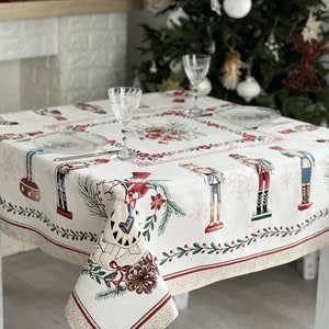 May include: A white tablecloth with a red and green border features a repeating pattern of nutcracker figurines, pine branches, and red poinsettia flowers.