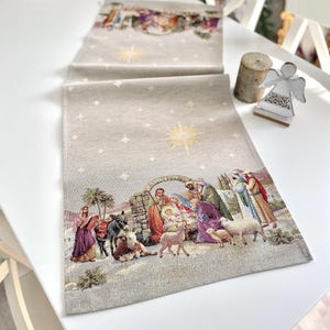 Op de afbeelding: Een beige tafelkleed met een geweven tapijtontwerp dat de kerststal voorstelt. De scène bevat Maria, Jozef, baby Jezus, een ezel, schapen en een ster. Het tafelkleed heeft een subtiele glans en is perfect om een ​​kerstachtige sfeer aan uw tafel toe te voegen.