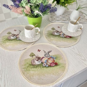 May include: Three beige placemats with a white border and an embroidered design of bunnies holding Easter eggs. The placemats are on a white table with a green plant and a white cup and saucer.