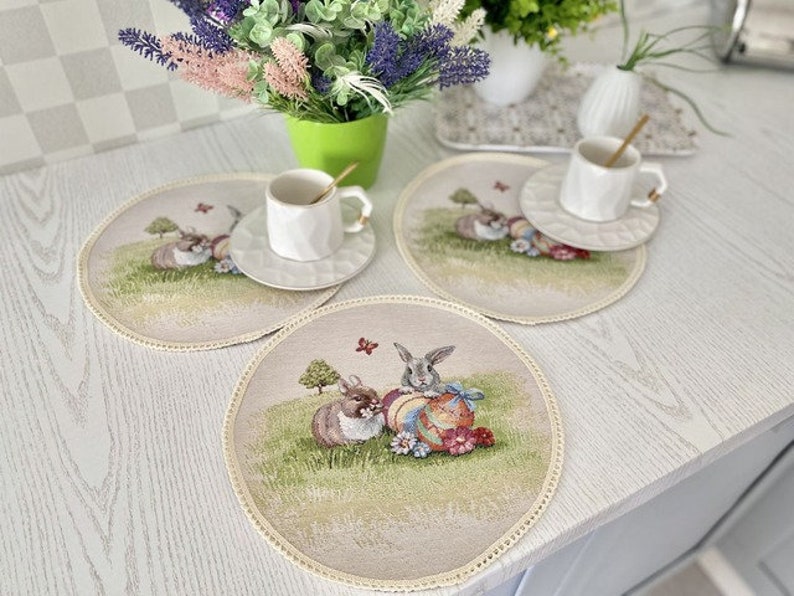 May include: Three beige placemats with a white border and an embroidered design of bunnies holding Easter eggs. The placemats are on a white table with a green plant and a white cup and saucer.