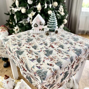 Puede incluir: Un mantel blanco con un diseño de pueblo de invierno con casas, árboles y personas. El mantel está sobre una mesa blanca con un árbol de Navidad en el fondo.
