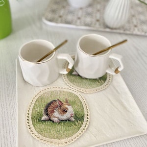 May include: Two white ceramic mugs with gold rims and handles, sitting on crocheted coasters with a bunny rabbit design. The coasters are on a white square tray.