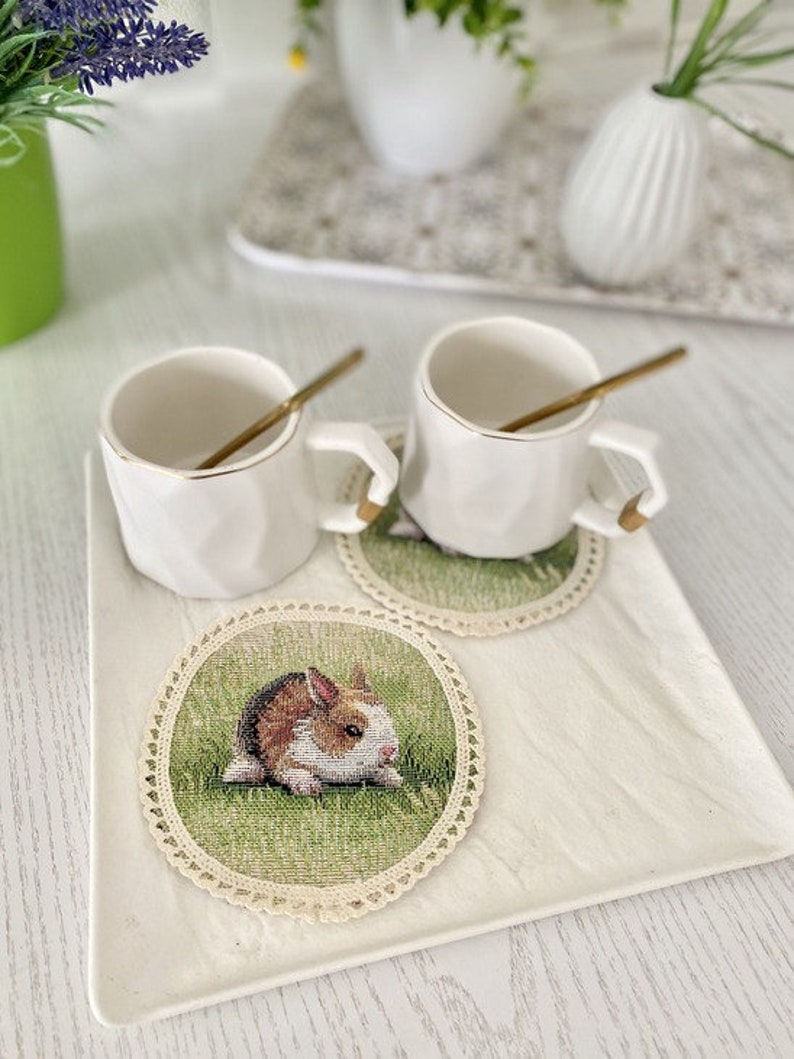 May include: Two white ceramic mugs with gold rims and handles, sitting on crocheted coasters with a bunny rabbit design. The coasters are on a white square tray.