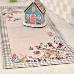 Op de afbeelding: Een lichtroze tafelkleed met een grijs-witte geruite rand. De loper heeft een geborduurd ontwerp met een konijn, bloemen, vlinders en paaseieren. Een blauw en wit peperkoekhuisje staat op de loper.